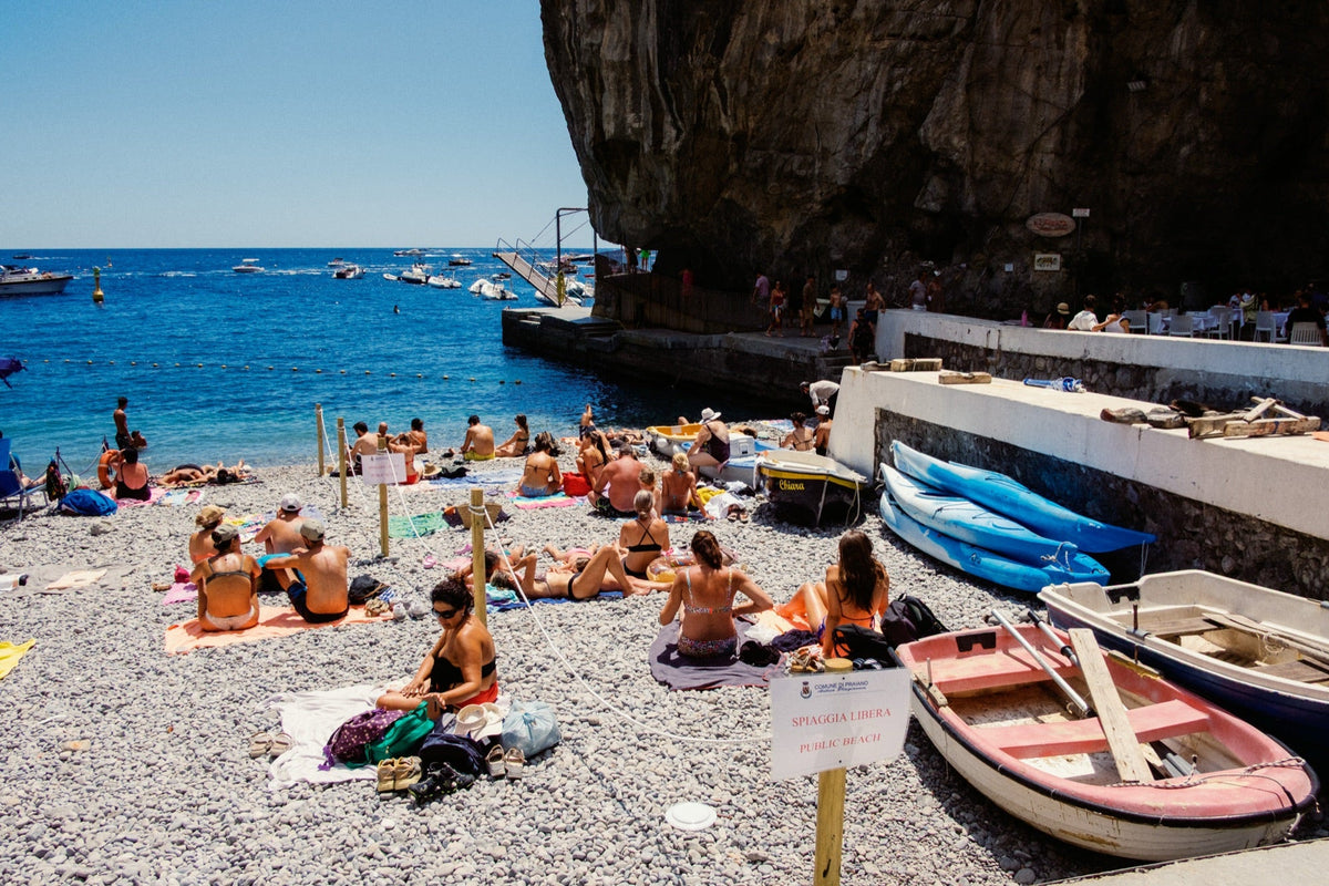 Sunbathers on a pebble beach below a rock overhang with moored rowing boats and kayaks, UltraMax 400 profile at +1 EV, shot on a Fujifilm X-Pro2